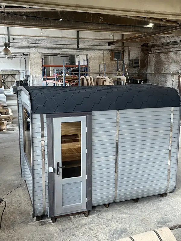 Vue d’angle extérieur d’un sauna cube 3 m Relax peint en gris clair, porte vitrée et banc latéral.