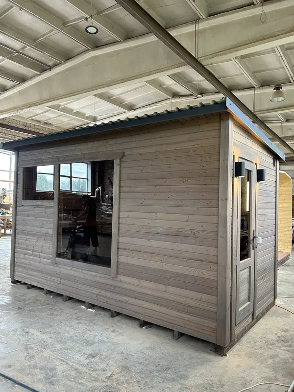 Vue d’un sauna box 4 m en bois clair/gris, pignon avec fenêtre et toit métallique, posé en hangar.