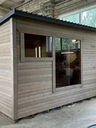 Sauna box en bois clair/gris avec grande fenêtre panoramique et fenêtre latérale, sous toit plat, en atelier.