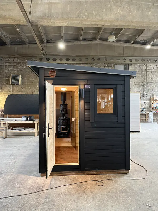 Vue d’entrée d’un sauna box noir avec porte ouverte, intérieur bois clair apparent, en atelier.