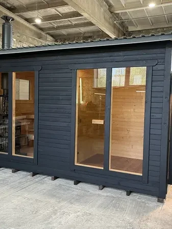 Sauna box foncé en ThermoWood avec deux larges baies vitrées et toit métallique, en hangar.