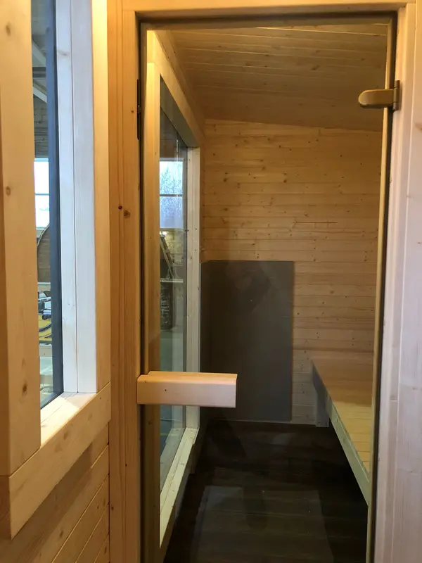 Couloir d’accès intérieur d’un sauna lambrissé clair, poignée de porte et lucarne verticale.