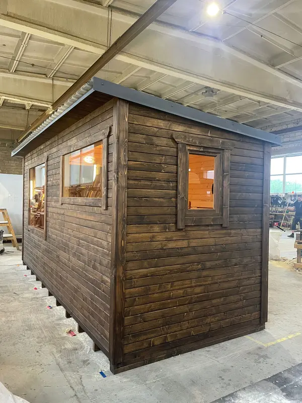 Sauna modulable en ThermoWood foncé, vue d’angle montrant une grande fenêtre et petite ouverture latérale sous un toit plat, en atelier.