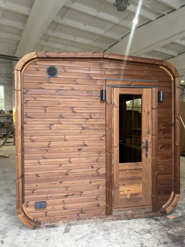 Pignon d’un sauna cube 5 m en ThermoWood brun, porte extérieure en bois et orifices de ventilation rondes.