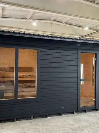 Sauna box 5 m peint en noir, façade avec grande fenêtre panoramique et porte vitrée décalée, en hangar.