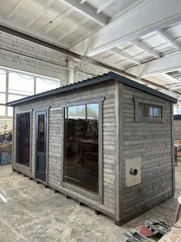 Vue d’angle d’un sauna box 5 m en ThermoWood gris clair, avec porte vitrée et grandes fenêtres latérales, posé en atelier.