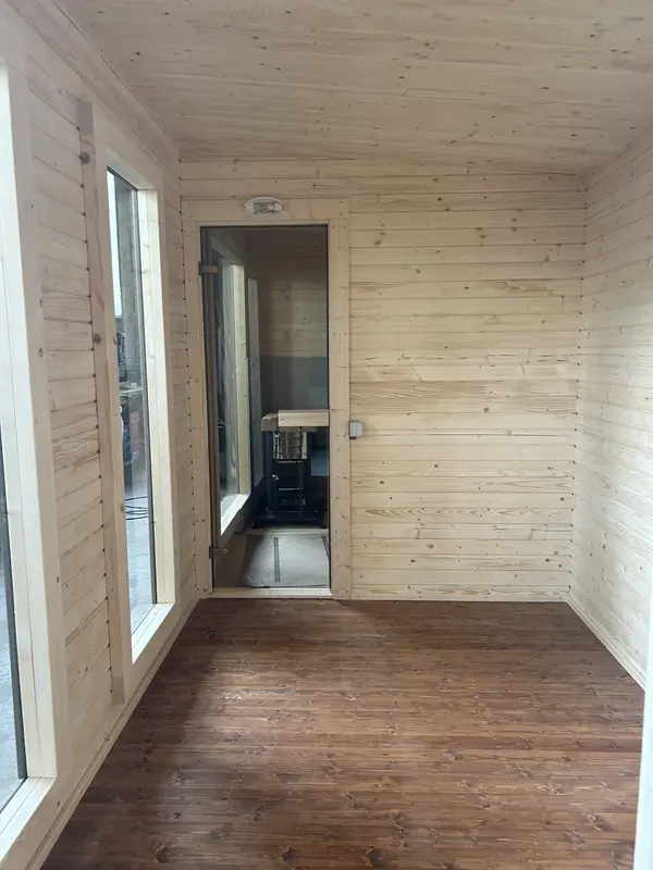 Couloir d’accès intérieur d’un sauna box 5 m, murs lambrissés clairs, sol en lattes de bois foncé et poêle visible.
