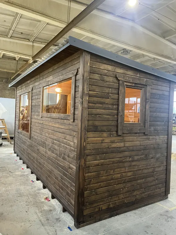 Angle latéral d’un sauna box 5 m ThermoWood brun foncé, grande fenêtre et petite ouverture sous toit plat.