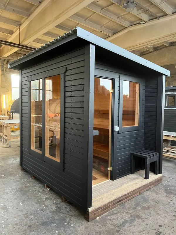 Sauna « L » extérieur peint en noir, porte vitrée et fenêtre latérale, toiture légèrement en pente.