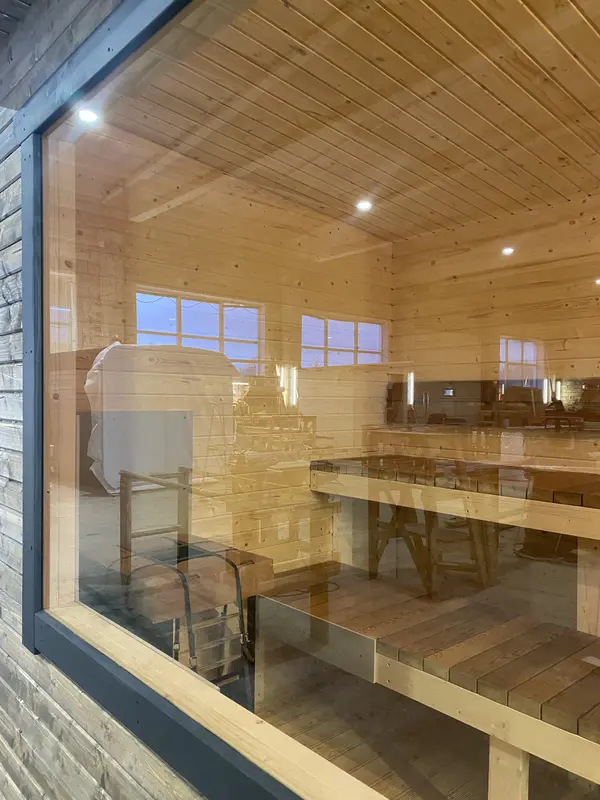 Intérieur d’un sauna en bois clair vu à travers la vitre, banquettes sur deux niveaux et plafond éclairé.