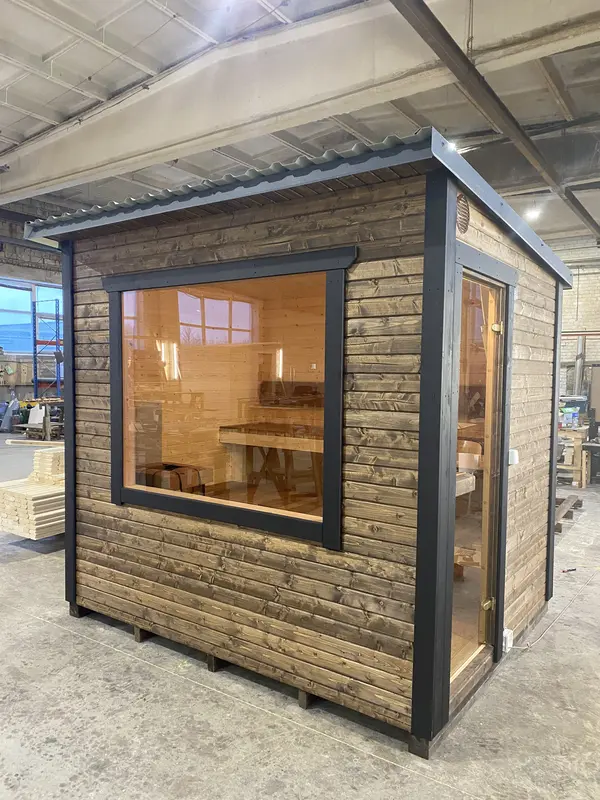 Vue d’angle d’un sauna box ThermoWood foncé avec grande fenêtre, intérieur lumineux visible.