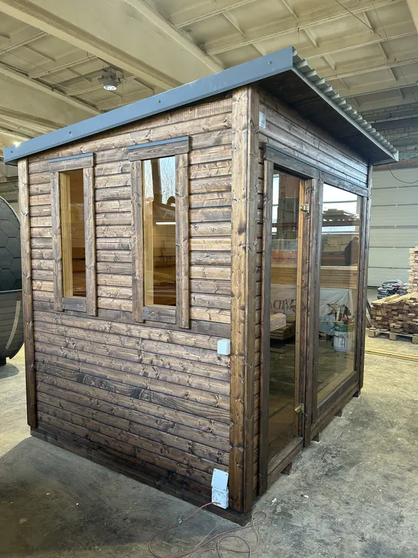 Sauna modèle « L » bardé en bois foncé vieilli, deux fenêtres verticales et porte vitrée, posé en intérieur