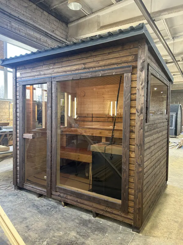 Sauna box ThermoWood foncé vue trois quarts, double porte vitrée et fenêtre latérale, en hangar.