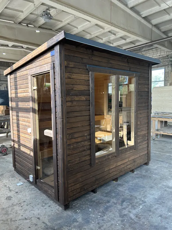 Autre angle d’un sauna « L » ThermoWood foncé, grande baie vitrée et prise d’air sur le côté.