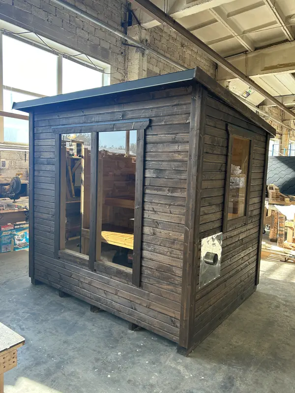 Sauna « L » tout en bois foncé avec double vitrage sur la face avant et toiture en tôle, en atelier.