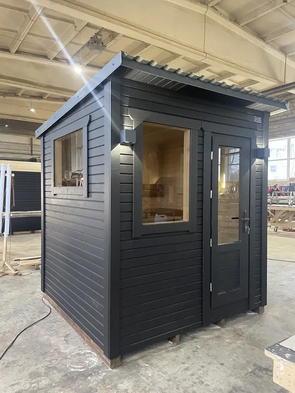 Sauna box ThermoWood peint en noir, porte vitrée ouverte et fenêtre latérale, en intérieur d’atelier.