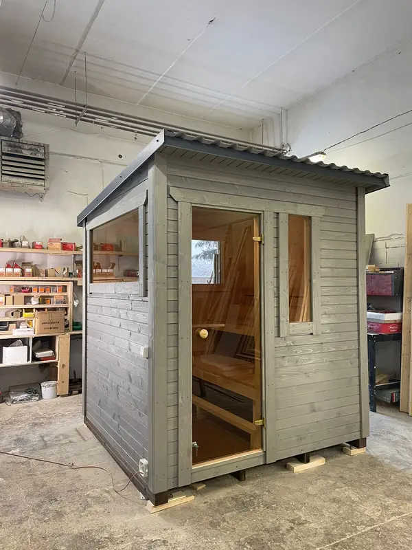 Sauna box modulable peint en gris clair, porte vitrée ouverte et fenêtre haute, en atelier.