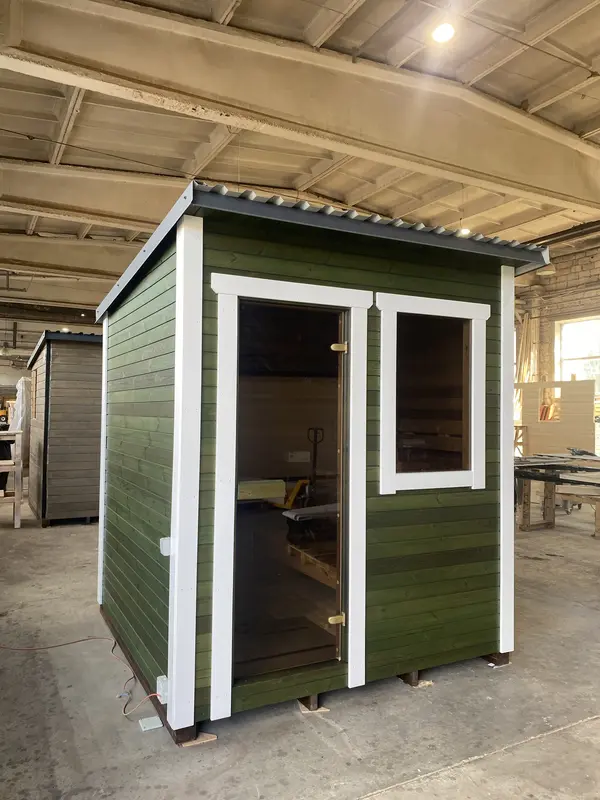 Sauna box peint en vert foncé avec encadrements blancs, porte pleine et châssis latéral vitré, en atelier.
