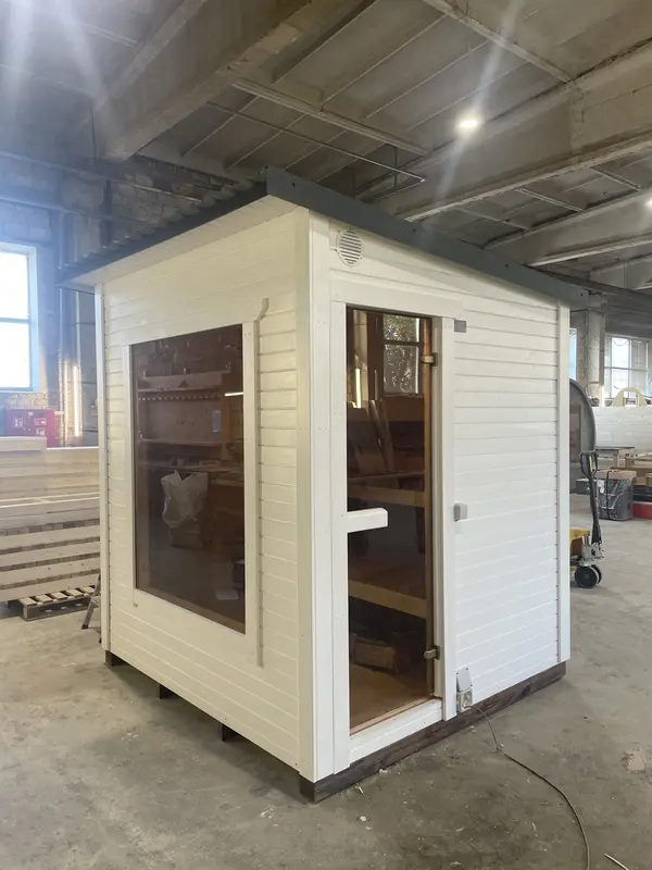 Sauna box modulable peint en blanc avec porte vitrée ouverte, intérieur bois visible, en atelier.
