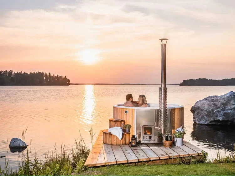Un couple se détend dans un bain nordique en bois rond avec un poêle à bois, situé sur une terrasse en bois au bord d'un lac, avec un coucher de soleil à l'horizon.