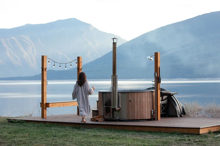 Une femme en robe de chambre marche vers un bain nordique en bois installé sur une terrasse en bois, face à un lac calme entouré de montagnes. Le bain nordique est équipé d'un poêle à bois.