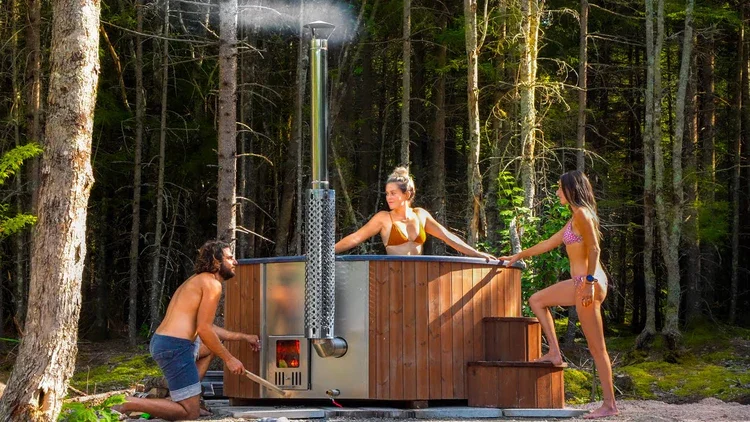 Trois personnes, en maillot de bain, se détendent autour d'un bain nordique en bois avec poêle à bois dans une forêt. L'une des personnes ajuste le feu pendant que les deux autres sont dans l'eau chaude